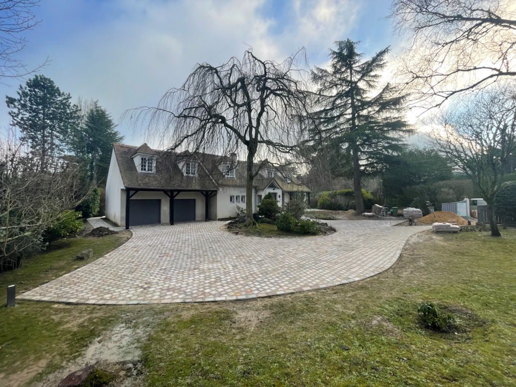 Une maison blanche avec une allée circulaire pavée, un grand arbre pleureur et un grand conifère dans un jardin gazonné.