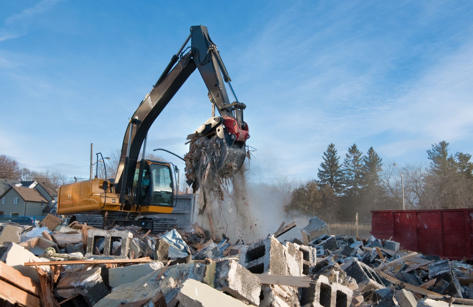 Une excavatrice jaune utilise une pince mécanique pour déplacer les débris de construction et les blocs de béton sur un chantier de démolition.