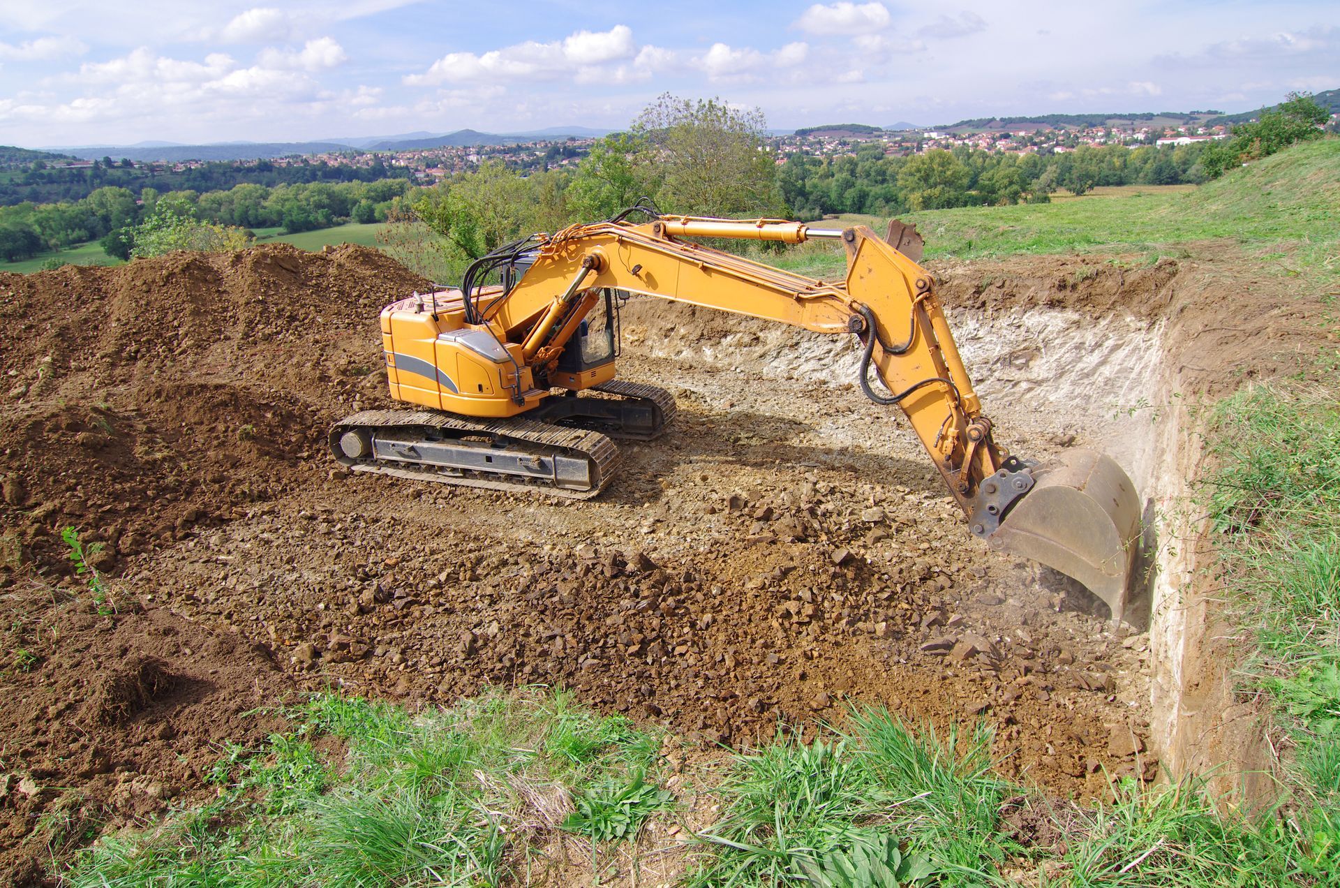 Une pelleteuse jaune creuse un grand trou dans une colline herbeuse sous un ciel bleu.