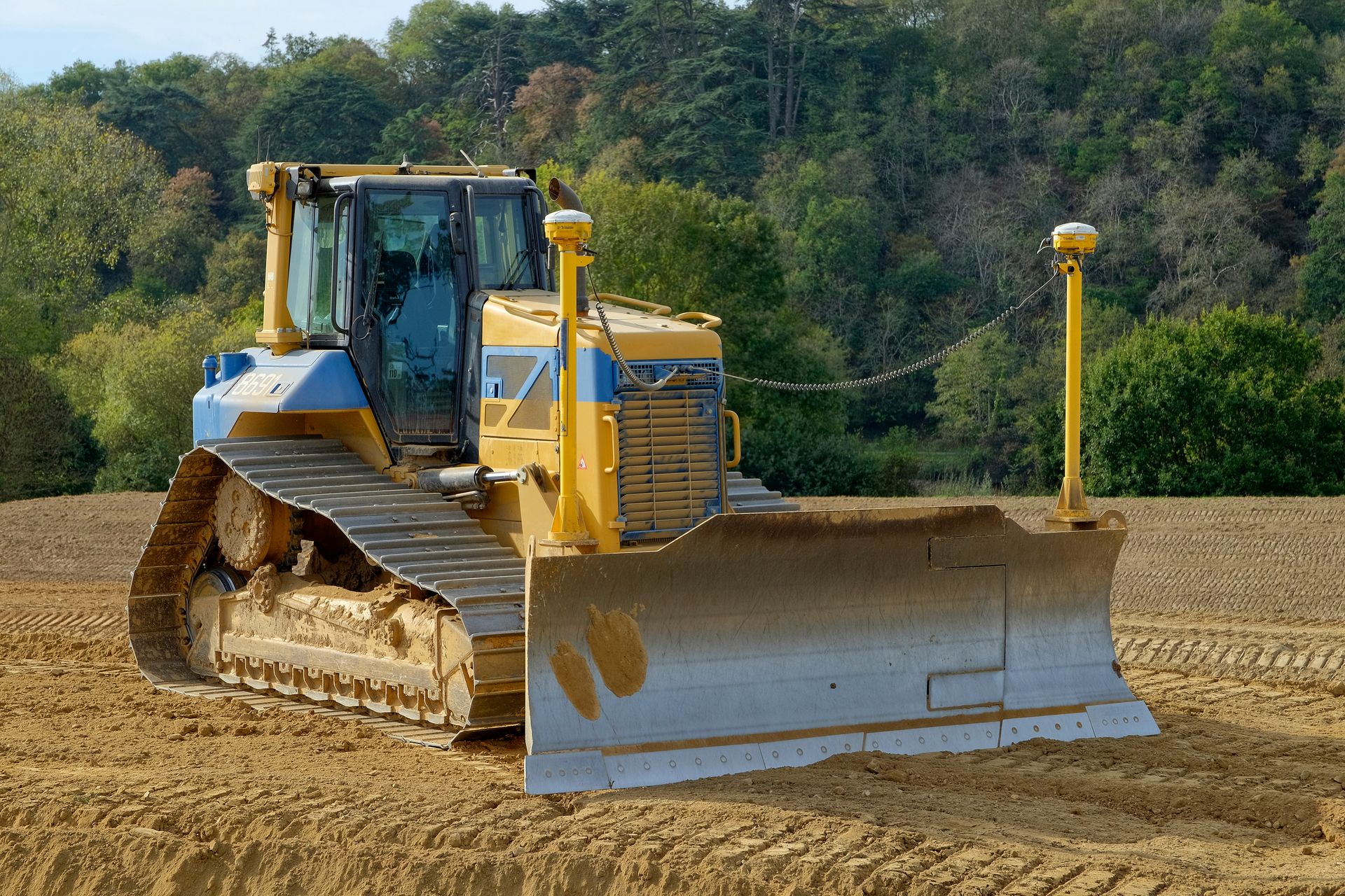 Un bulldozer jaune et bleu, équipé d'antennes GPS sur sa lame, est stationné sur un chantier de construction en terre battue, avec une forêt en arrière-plan.