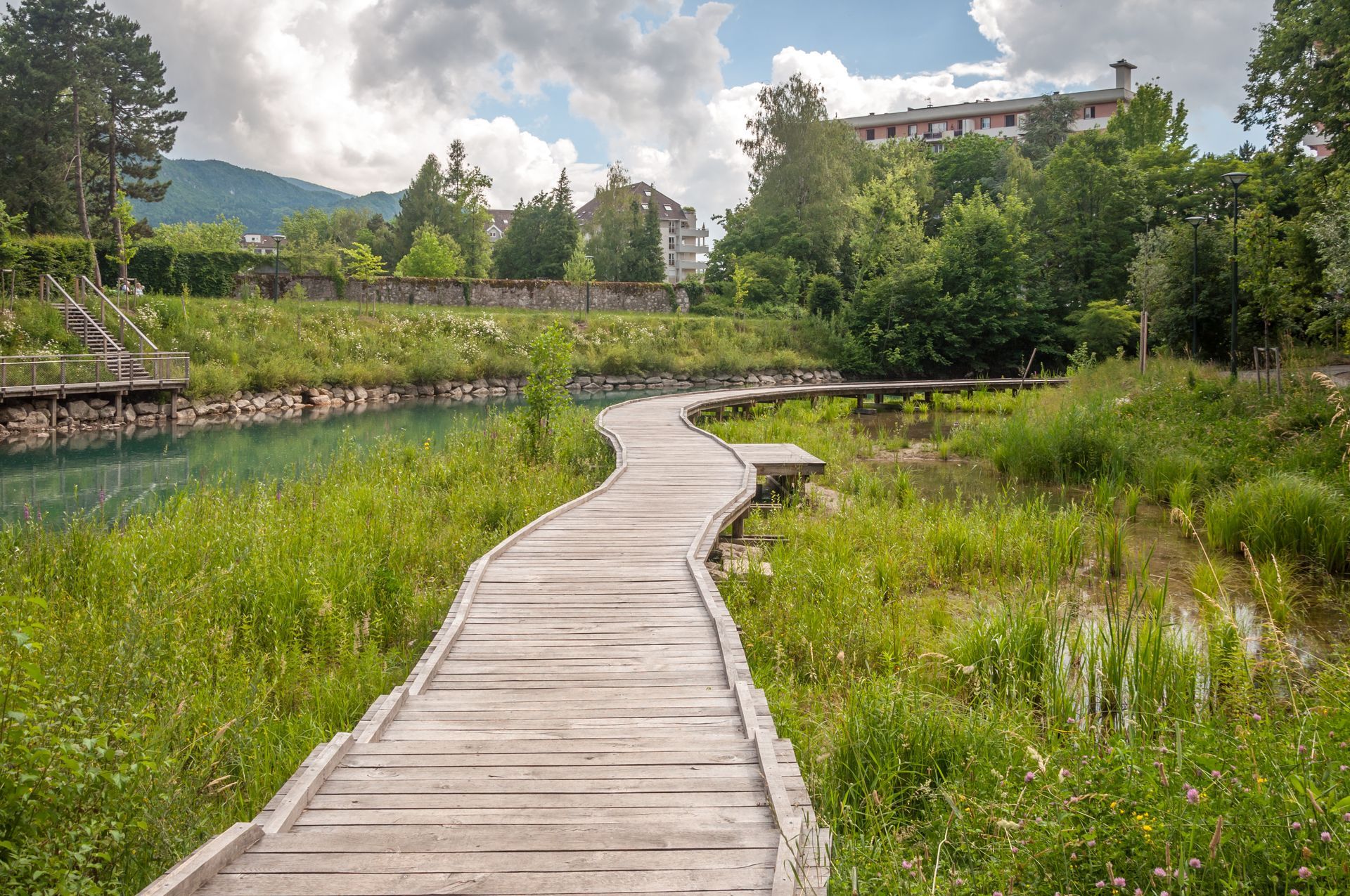 Une passerelle en bois serpente à travers des zones humides verdoyantes jusqu'à un petit étang et des bâtiments au loin, sous un ciel nuageux.