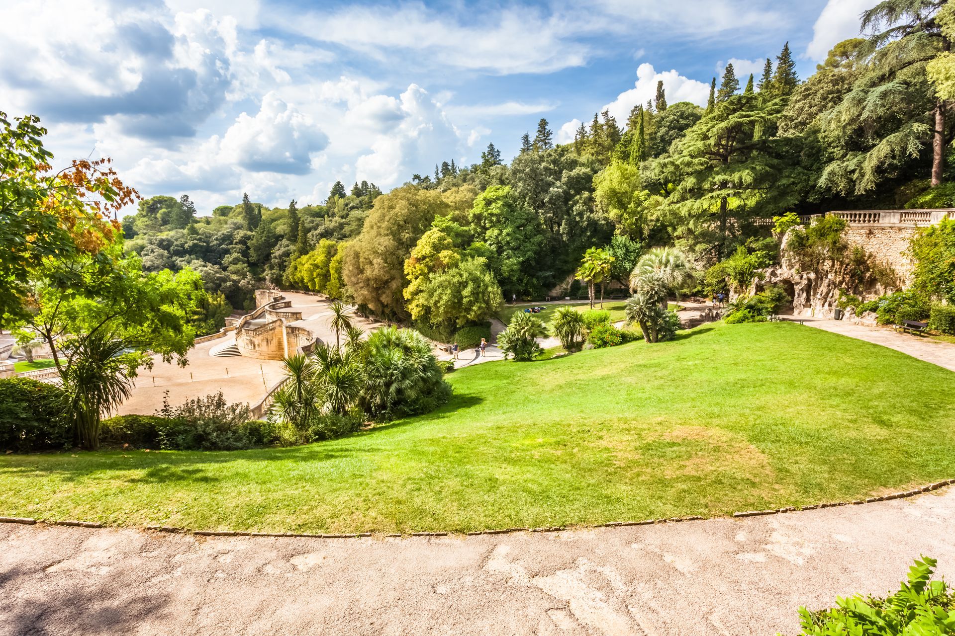 Vue panoramique d'un parc public ensoleillé avec des pelouses verdoyantes, un chemin pavé et des arbres denses et luxuriants sous un ciel bleu nuageux.