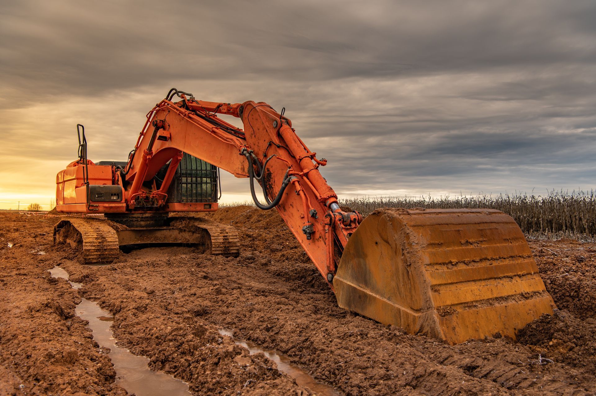 Une pelleteuse orange se dresse sur un chantier boueux et couvert, son grand godet posé à même le sol.