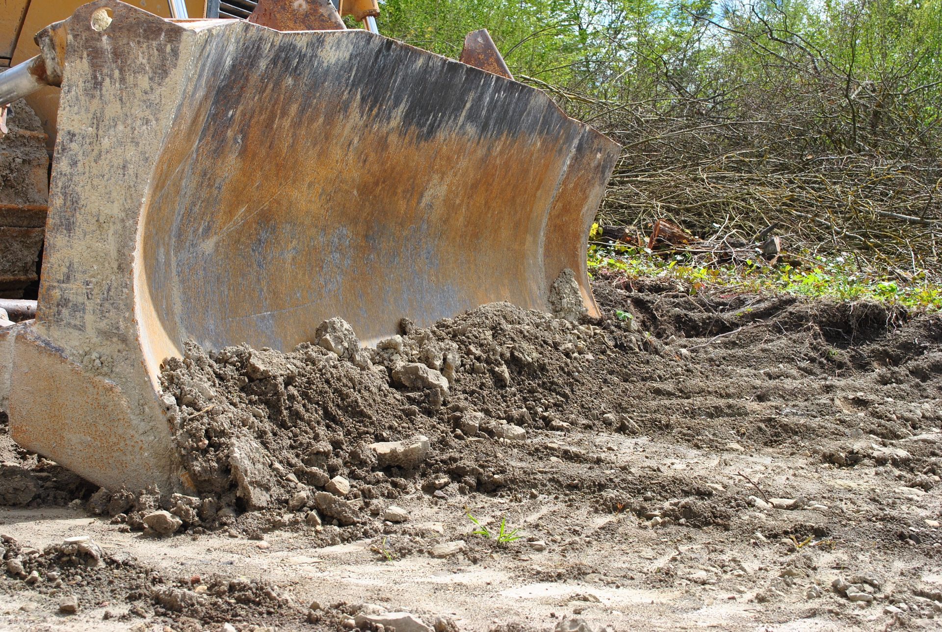 La lame d'un bulldozer imposant pousse un tas de terre brune et de poussière sur un terrain extérieur où l'on aperçoit du feuillage vert en arrière-plan.