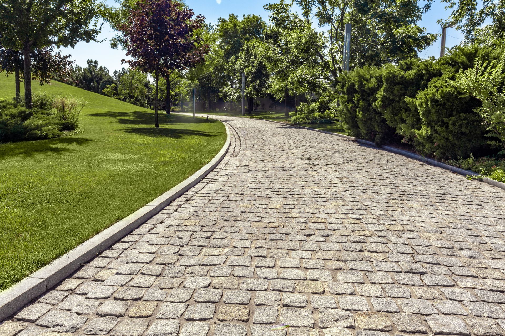 Un chemin pavé sinueux serpente à travers un parc d'un vert éclatant, avec une pelouse impeccablement entretenue, des arbres et des haies, sous un ciel bleu.