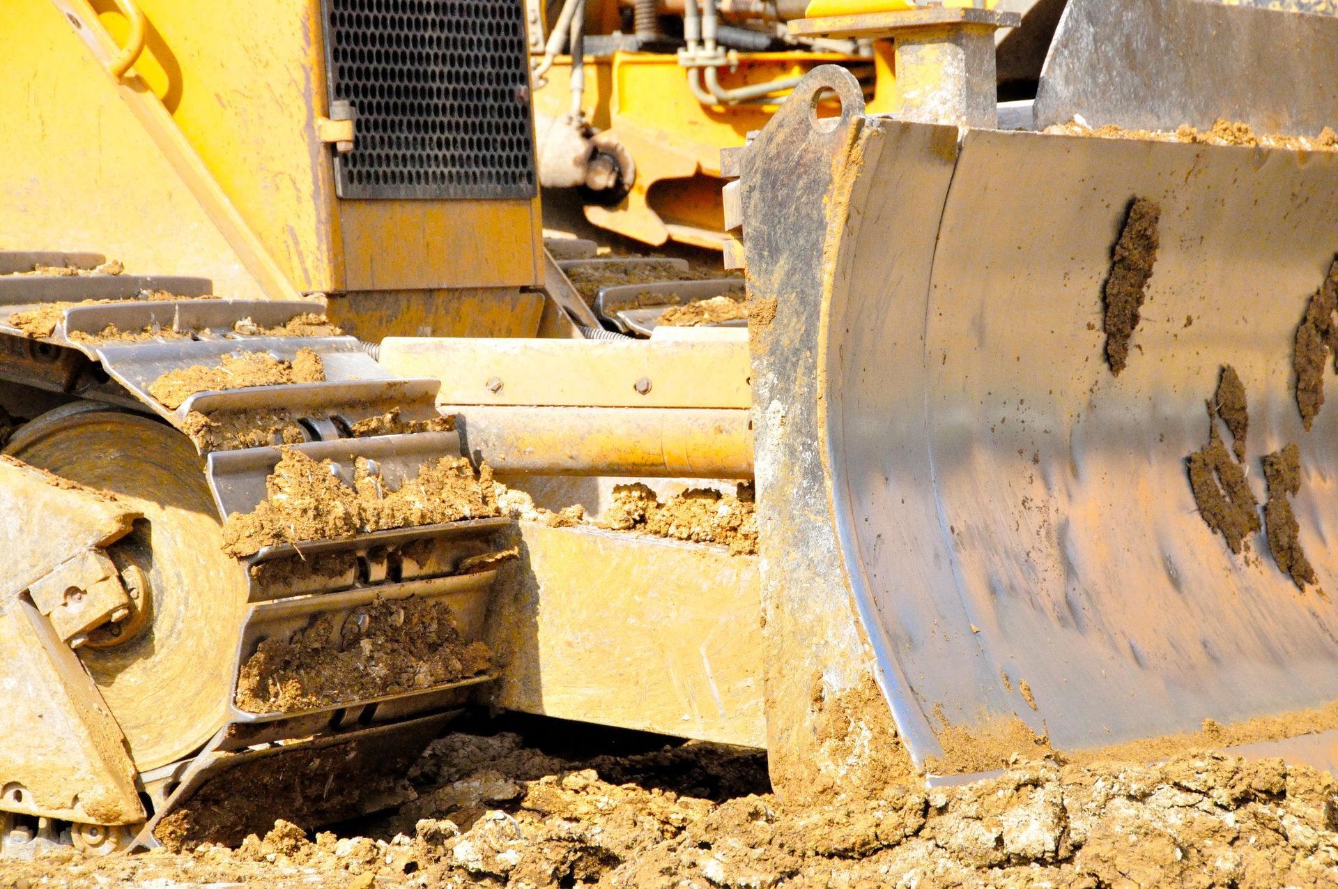 Gros plan sur la lame métallique et les chenilles d'un bulldozer jaune, recouvertes de boue, sur un chantier de construction.
