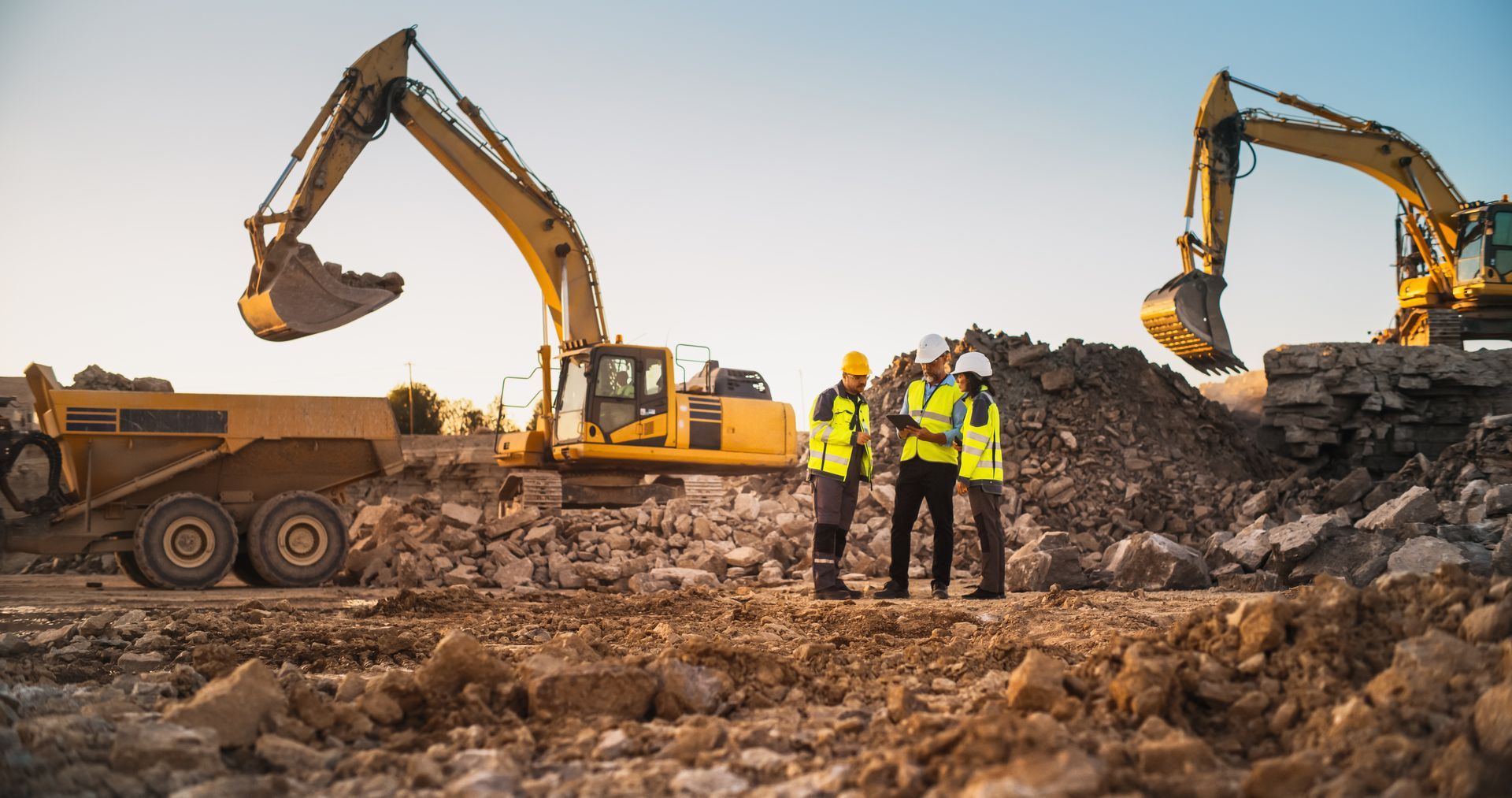 Trois ouvriers du bâtiment, casqués et vêtus de gilets de sécurité, examinent des plans sur un chantier où se trouvent deux excavatrices et un camion-benne.