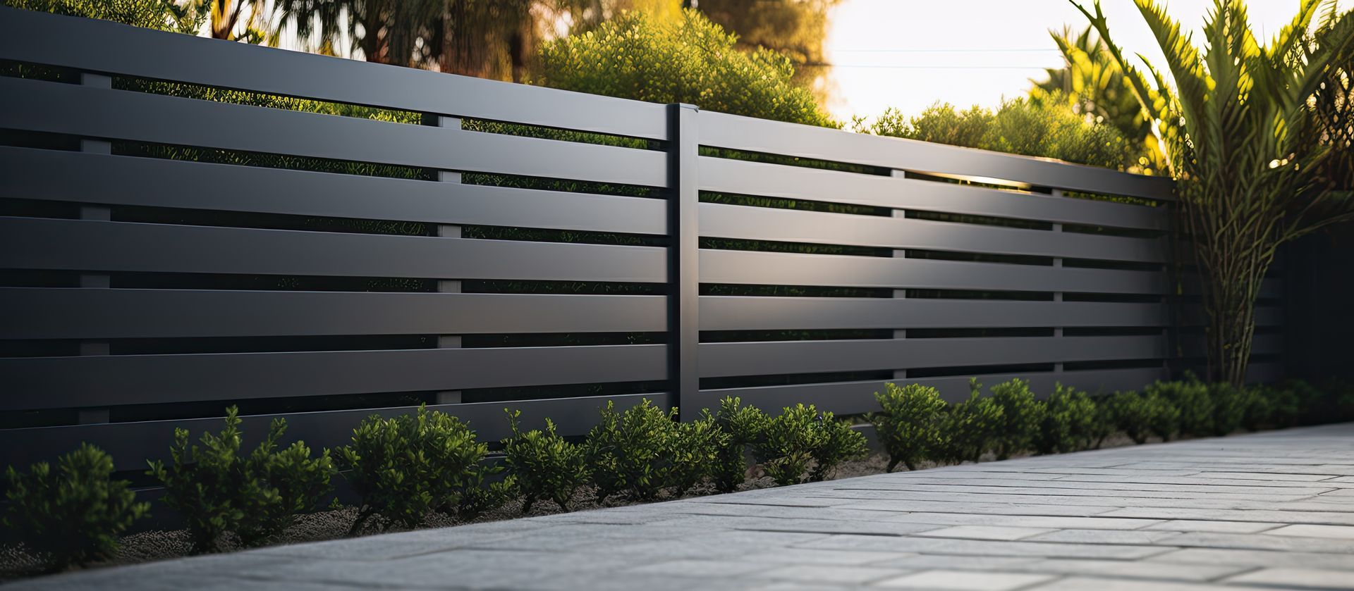 Clôture moderne à lattes horizontales gris foncé, avec une végétation luxuriante se détachant sur un chemin de pierre au coucher du soleil.