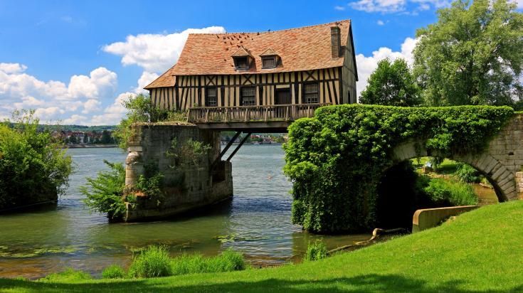 Une vieille maison à colombages construite au-dessus de l'eau, reliée à un pont de pierre recouvert de lierre vert par une journée ensoleillée.