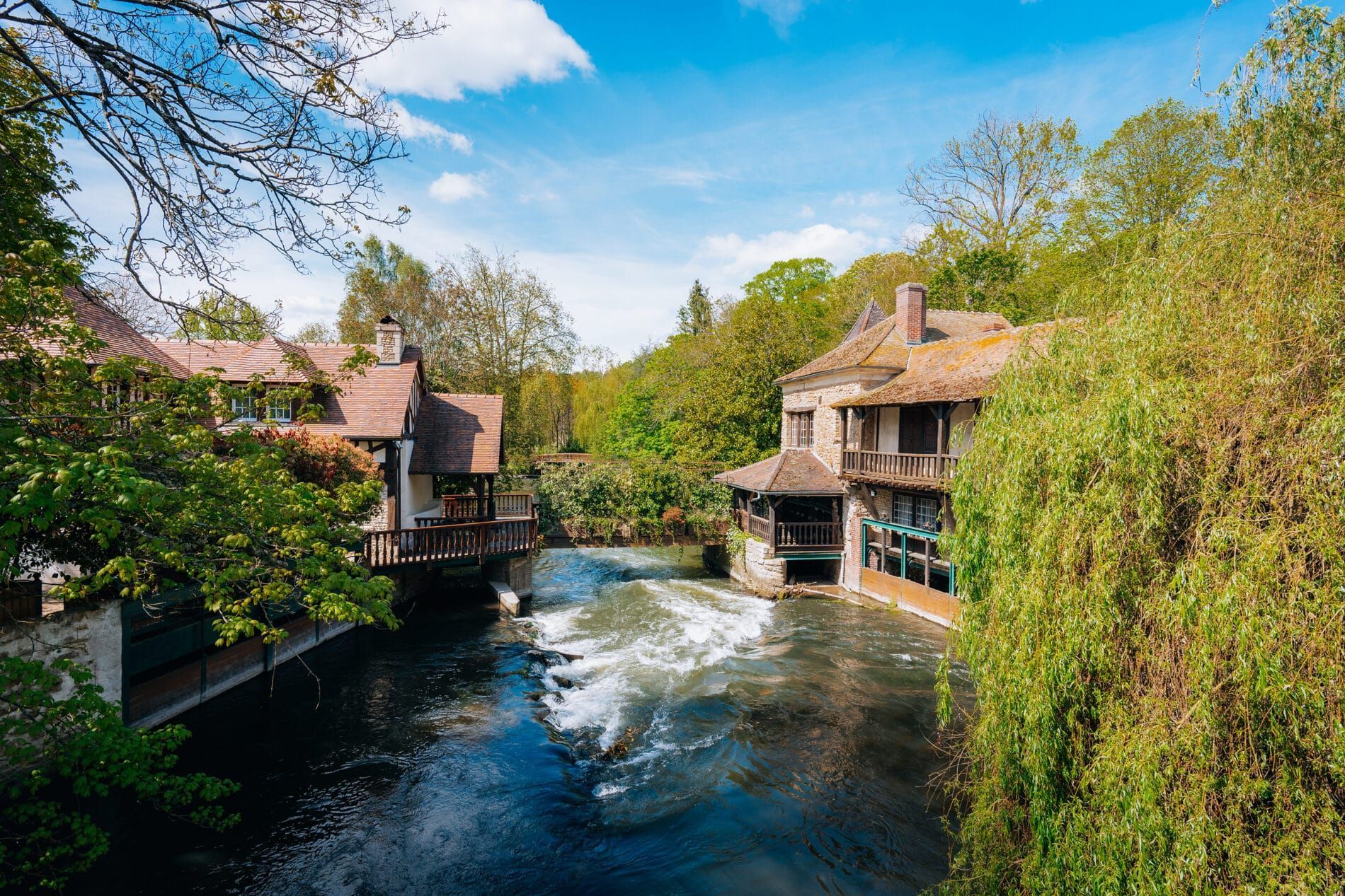 Un bâtiment en pierre au toit rustique se dresse au bord d'une rivière impétueuse, entouré d'une végétation luxuriante sous un ciel d'un bleu éclatant.