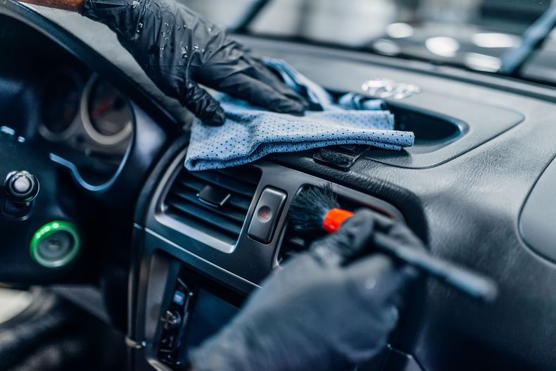 Deux mains gantées nettoient le tableau de bord d'une voiture avec une petite brosse et un chiffon en microfibre.