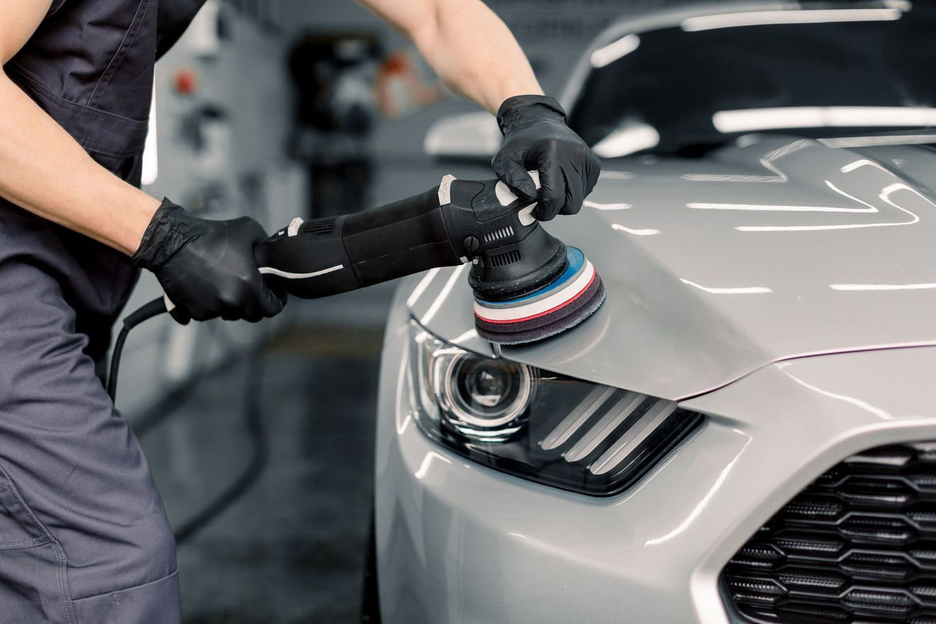 Une personne en tenue de travail et gants noirs utilise une polisseuse électrique sur le capot d'une voiture.