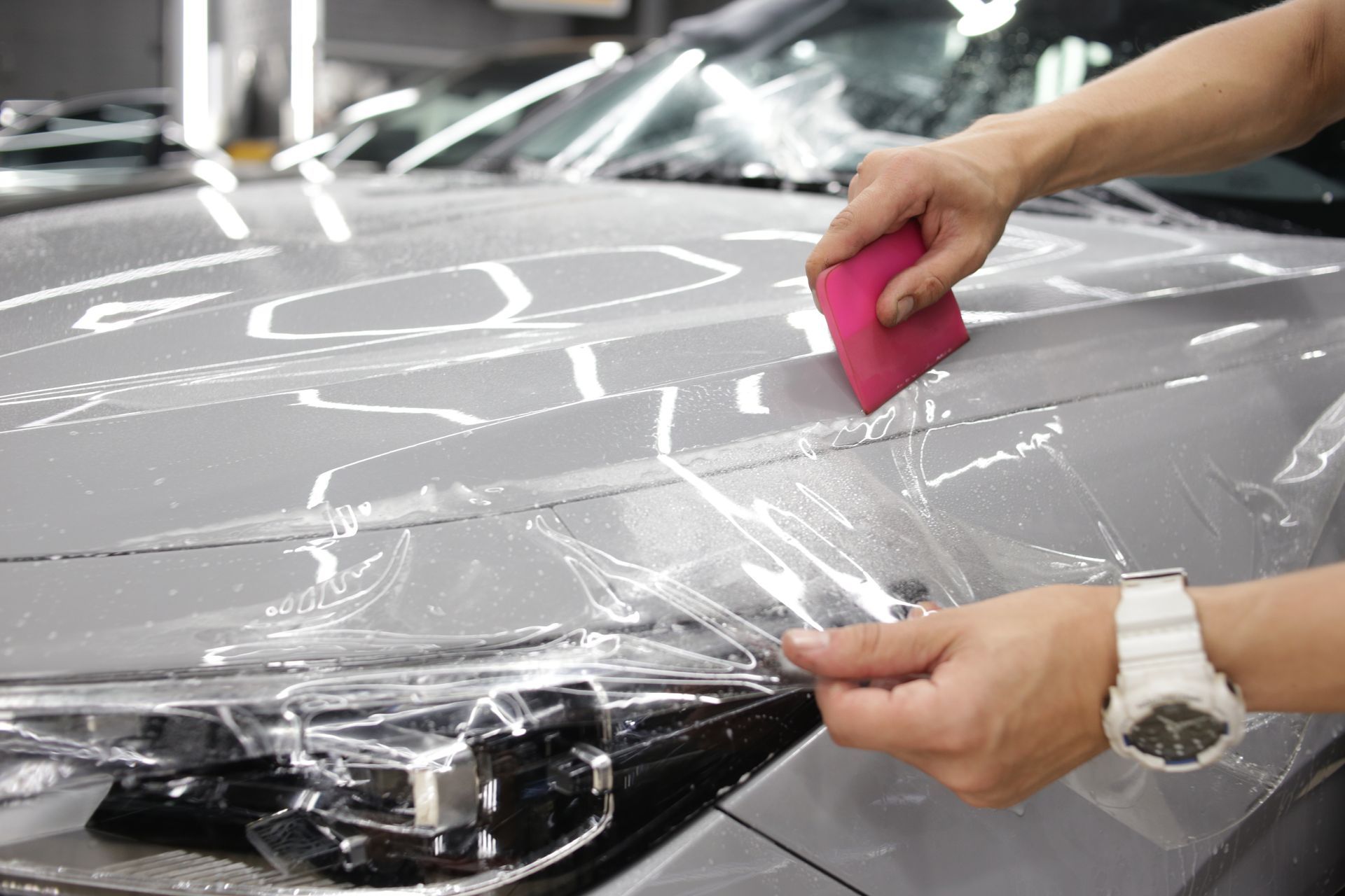 Une personne utilise une raclette rose pour appliquer un film protecteur transparent sur le capot gris d'une voiture dans un atelier.