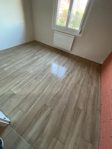 Pièce vide avec sol carrelé imitation bois, petite fenêtre et radiateur mural.