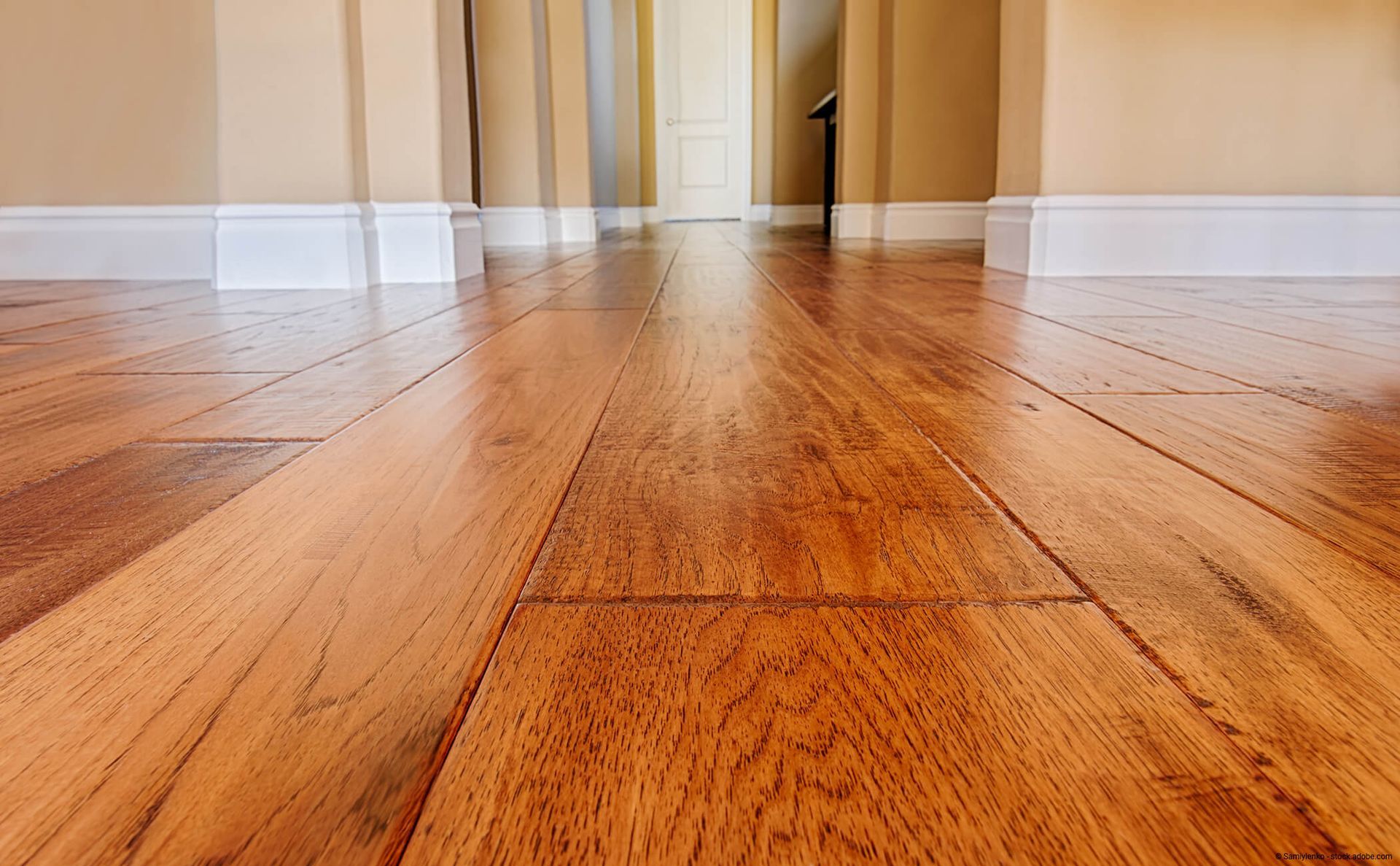 Aufnahme von warmfarbigem Hartholzboden in einem Flur mit weißen Fußleisten und einer Tür im Hintergrund.
