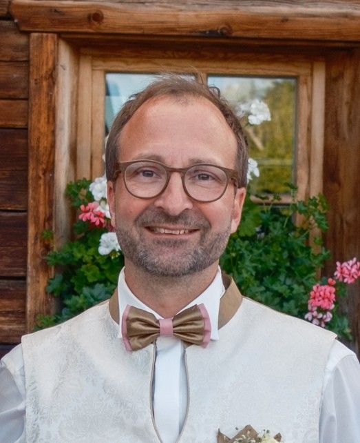 Eine Person in einer hellen Weste und einer gold-rosa Fliege posiert vor einem rustikalen Holzfenster mit Blumen.