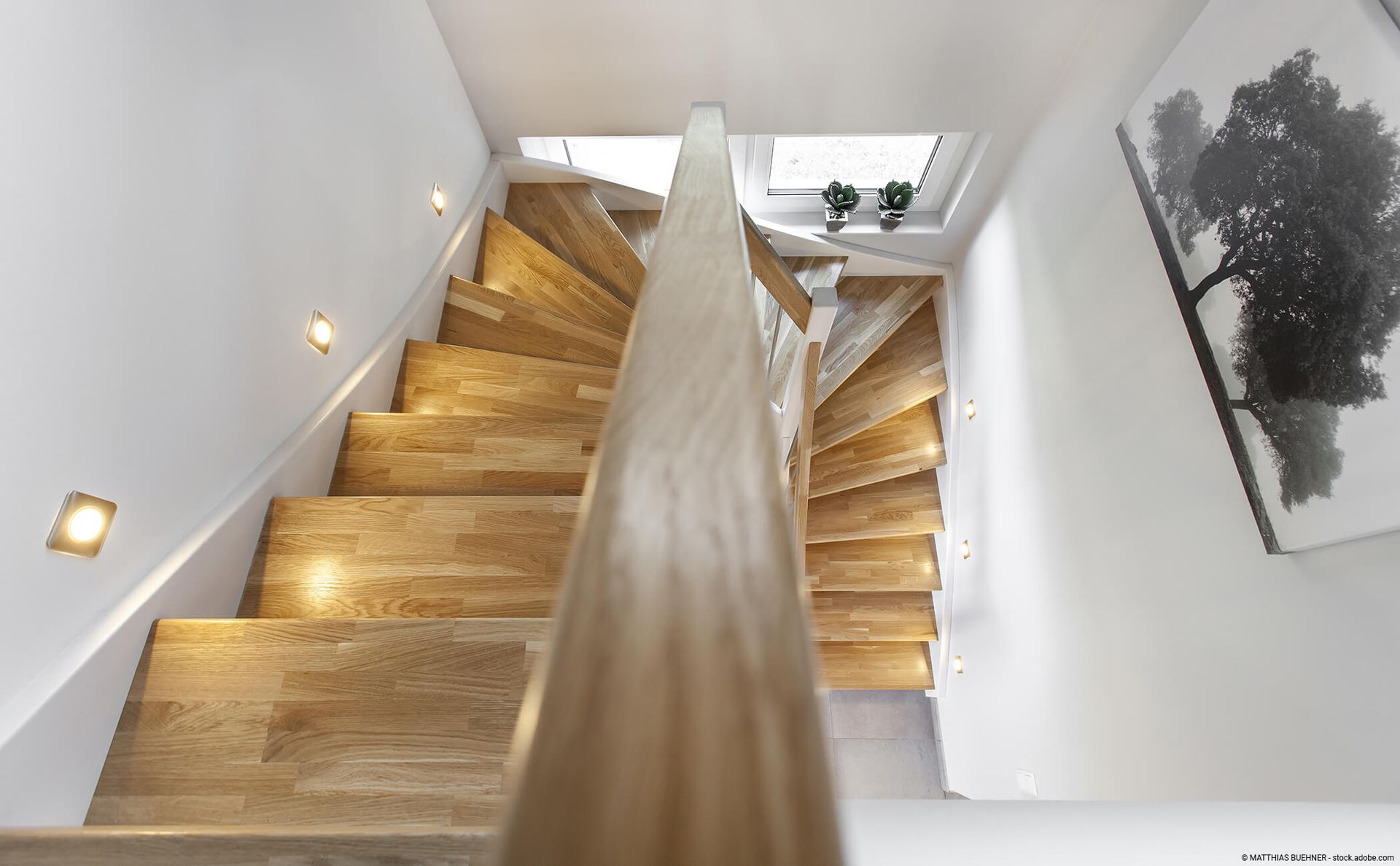 Ein Blick aus der Vogelperspektive auf eine Holztreppe mit warmer Stufenbeleuchtung und einem zentralen Handlauf