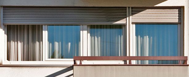 Vue extérieure d'un immeuble avec fenêtres, stores et rideaux. Une rambarde de balcon est visible au premier plan.