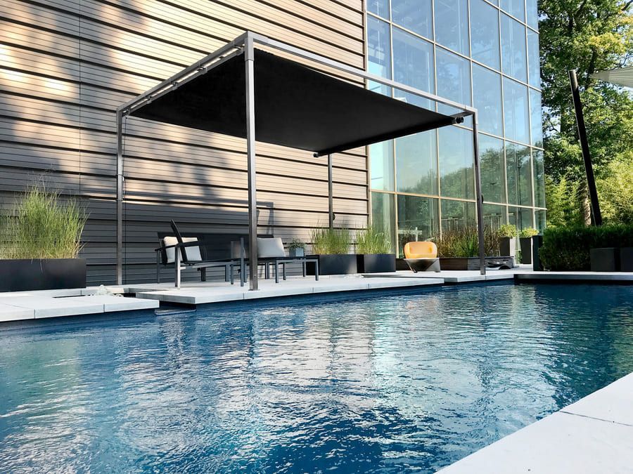 Piscine avec structure d'ombrage moderne et sièges devant un bâtiment aux baies vitrées.