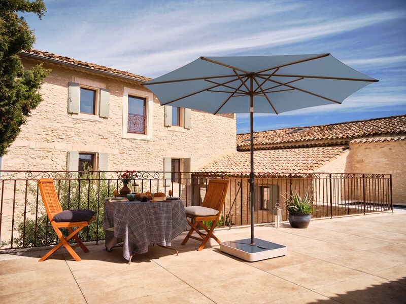 Terrasse avec table, chaises et parasol bleu, devant un bâtiment en pierre avec balcon.