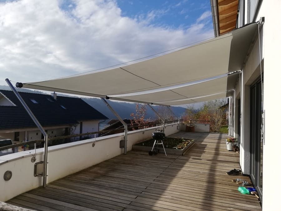 Une voile d'ombrage blanche protège une terrasse en bois. Supports en acier inoxydable fixés au bâtiment et à la rambarde.