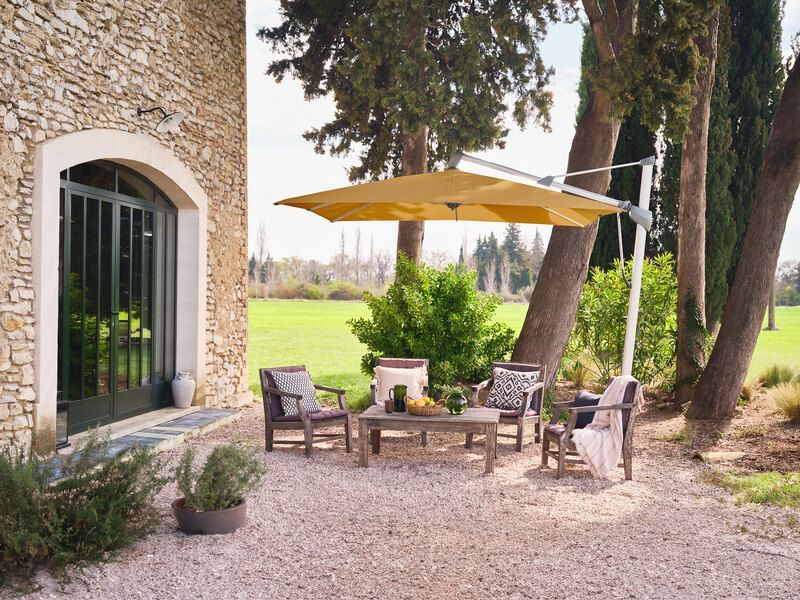 Terrasse extérieure avec parasol jaune déporté, bâtiment en pierre, arbres et gravier.