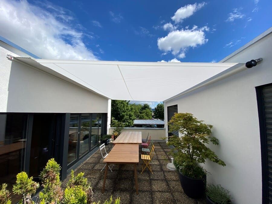 Par une journée ensoleillée, un parasol blanc recouvre une terrasse où se trouvent une table, des chaises et des plantes en pot.
