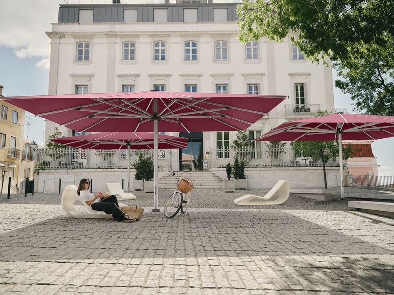 Une femme se détend sous un parasol rouge sur une terrasse en pierre avec des chaises longues blanches, un vélo et un bâtiment.