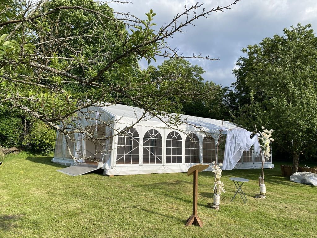 Une tente blanche est dressée sur une pelouse, avec des arbres en arrière-plan, probablement pour un mariage.