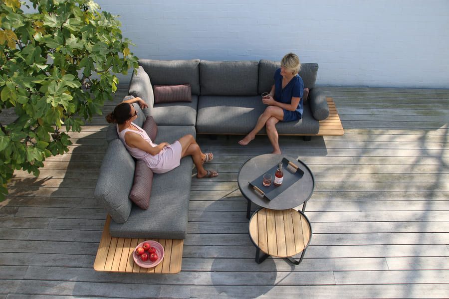 Deux femmes se détendent sur un canapé d'angle gris d'extérieur, sur une terrasse en bois. L'une est allongée, l'autre assise droite.