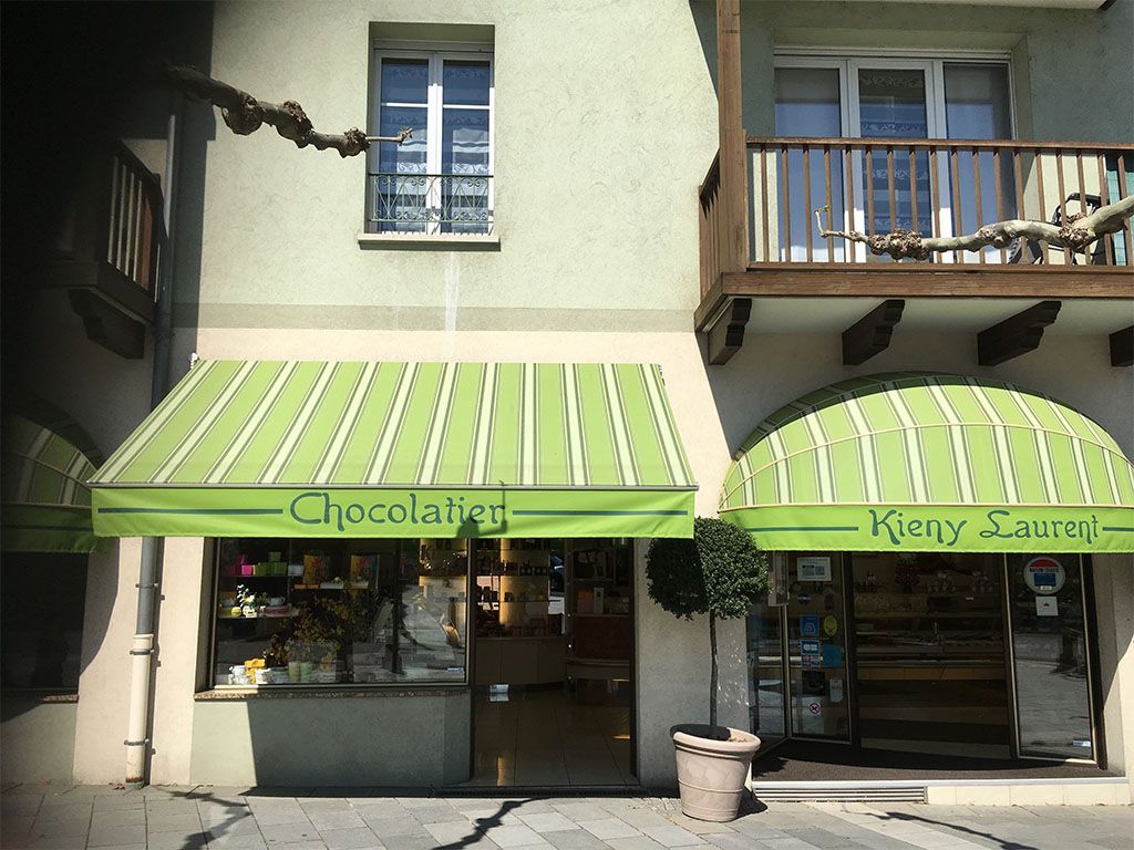 Boutiques Chocolatier et Kierry Saloni avec auvents verts. Façade d'immeuble vert clair, cadre extérieur.