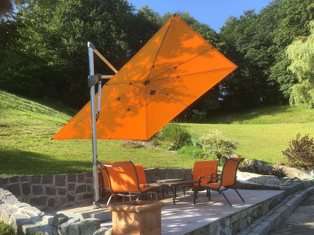 Un parasol orange ombrage un coin salon sur une terrasse en pierre ; de l'herbe verte et des arbres en arrière-plan.