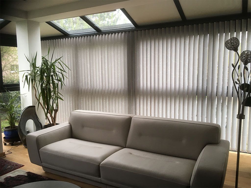 Salon avec un canapé beige, des stores verticaux et une grande plante.