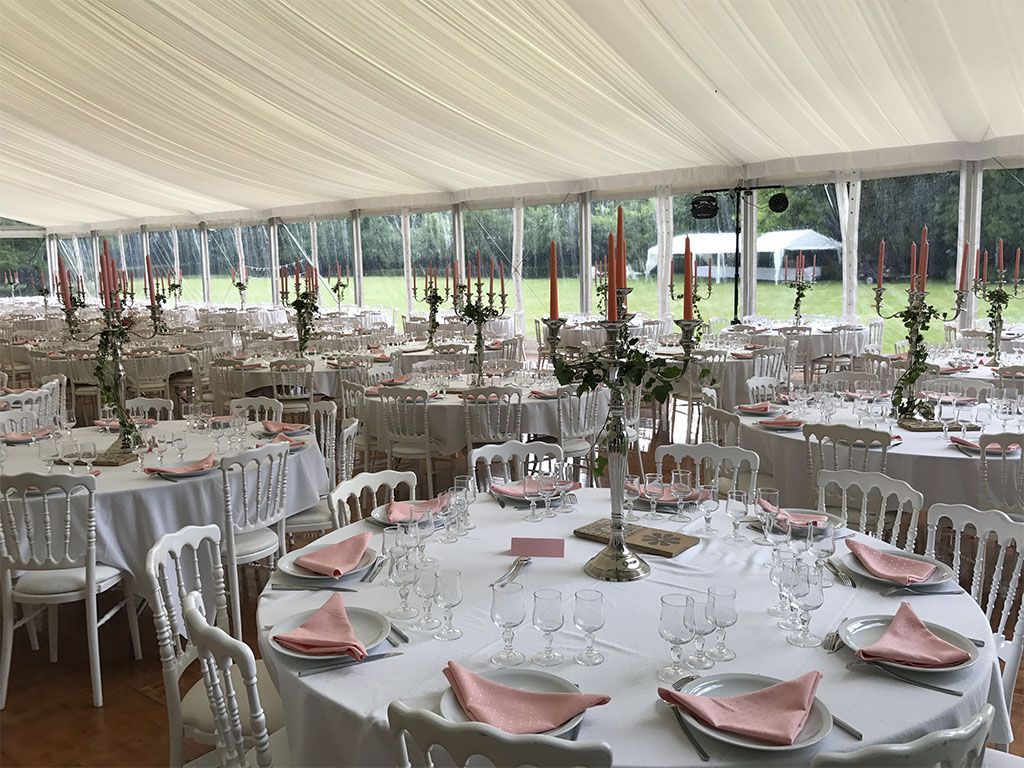Sous une tente blanche, des tables rondes sont dressées pour un événement formel, avec des chaises blanches, des serviettes roses et des candélabres.