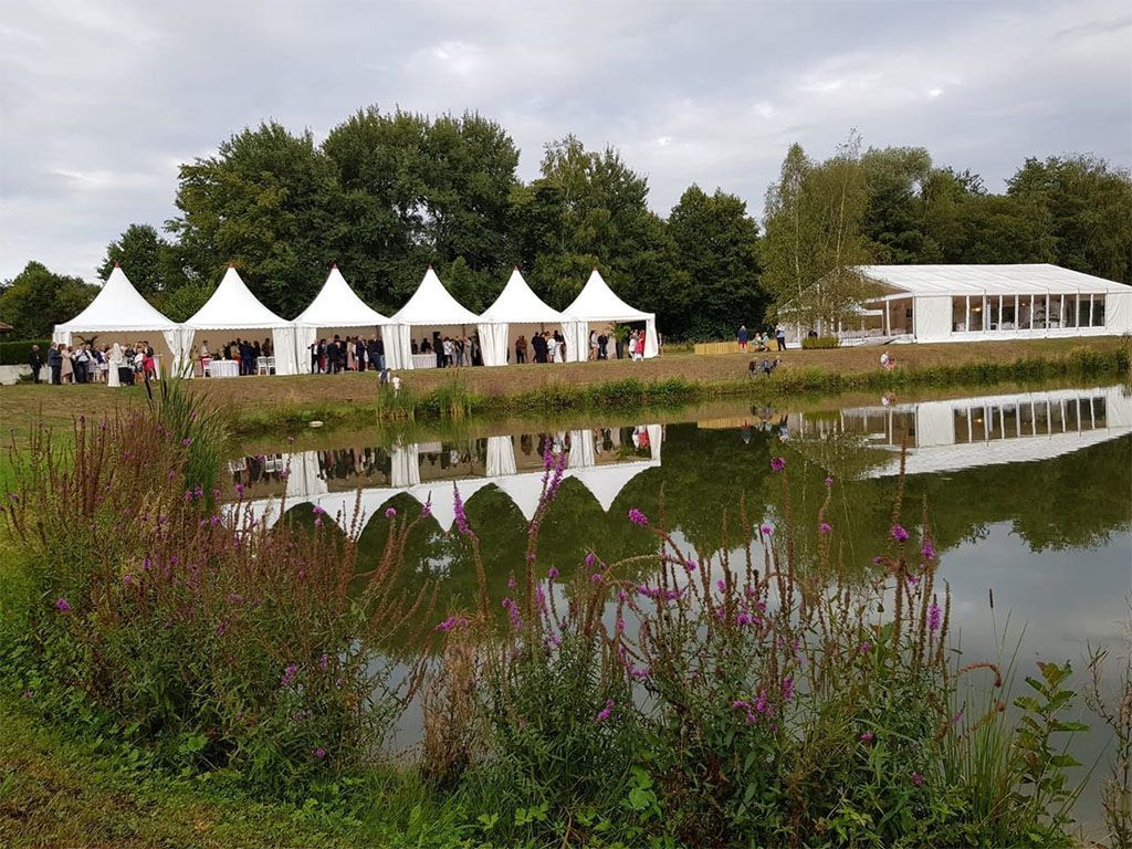Événement sous chapiteau au bord d'un étang. Reflets des tentes et d'un bâtiment dans l'eau, avec des arbres verts et des fleurs violettes.