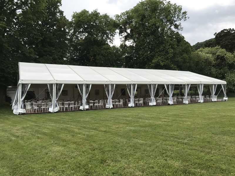 Longue tente blanche dressée sur une pelouse verte, avec tables et chaises pour une réception. Arbres en arrière-plan.