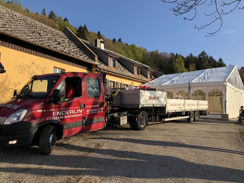 Un camion rouge avec remorque est stationné près d'un bâtiment et d'une tente blanche, probablement pour un événement.
