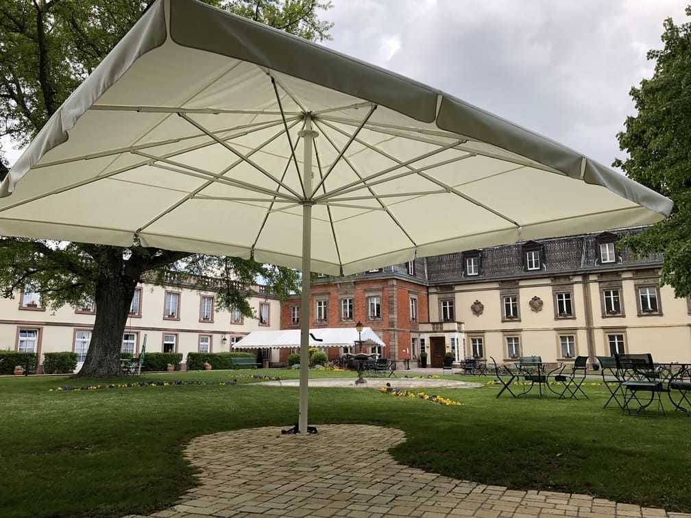 Grand parasol carré au-dessus d'une terrasse en briques devant un bâtiment de style européen, sur une pelouse.