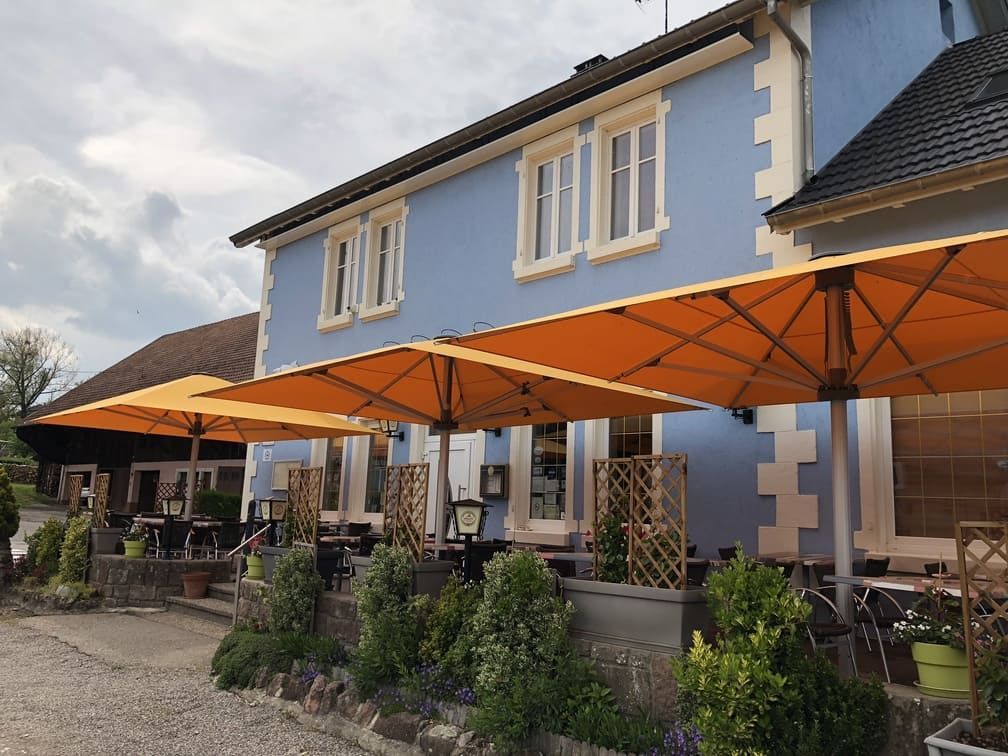 Restaurant avec terrasse sous des parasols orange. Bâtiment bleu, végétation verte. Ciel nuageux.