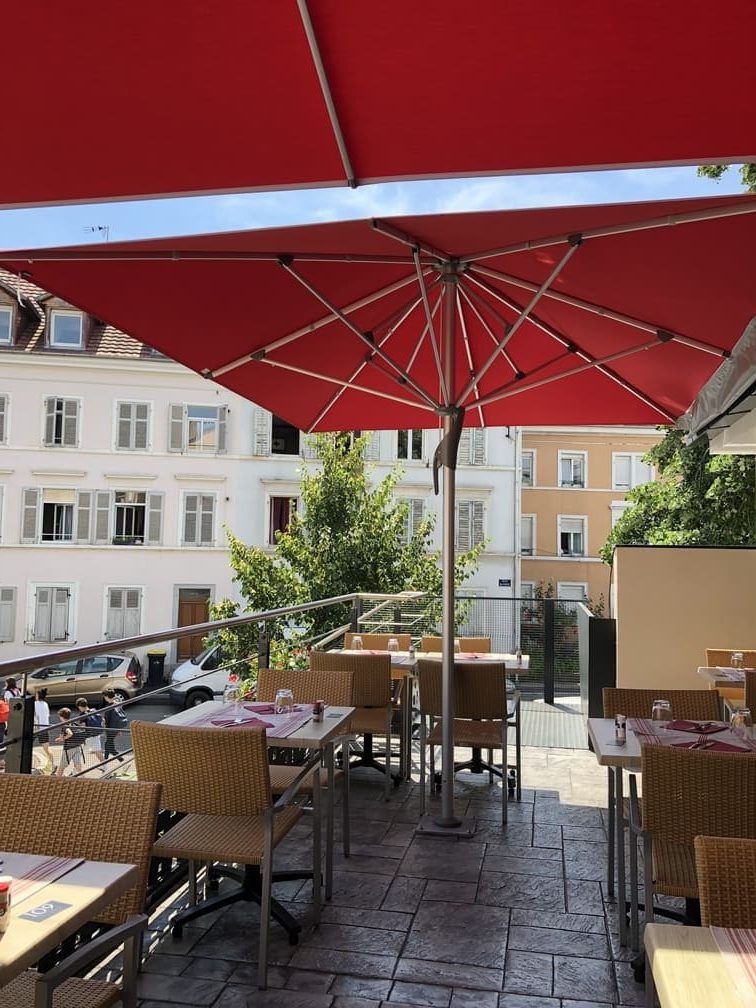 Terrasse de restaurant avec parasols rouges, tables et vue sur des bâtiments blancs et des arbres.