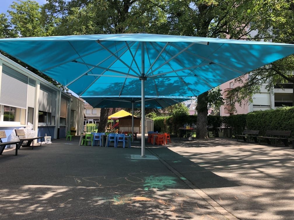 Grande structure bleue ombragée au-dessus d'une cour d'école avec des tables, des chaises et des arbres.