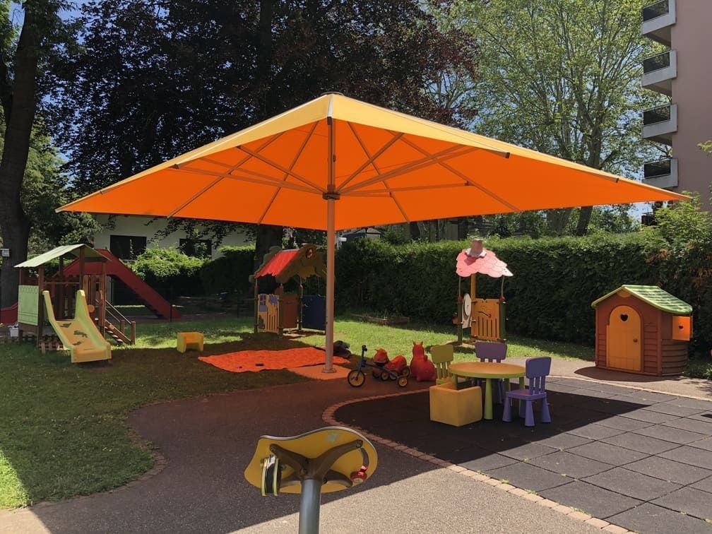 Un grand parasol orange protège du soleil une aire de jeux avec des toboggans, des maisonnettes et du mobilier à la taille des enfants.
