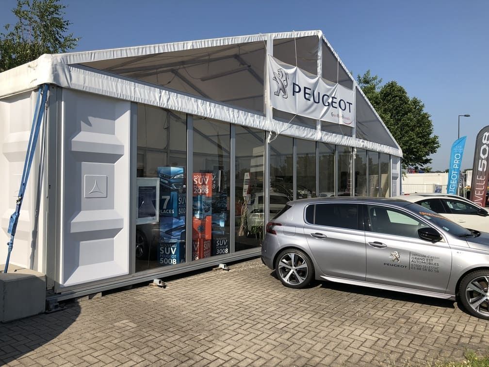 Boutique éphémère Peugeot avec vitrines, une voiture argentée garée à l'extérieur. Structure de tente blanche, logo Peugeot visible.