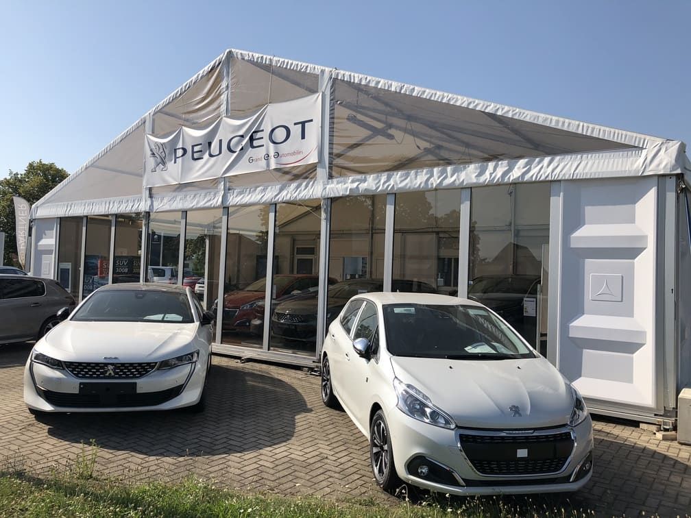 Deux voitures Peugeot blanches garées devant une tente de concessionnaire Peugeot par une journée ensoleillée.