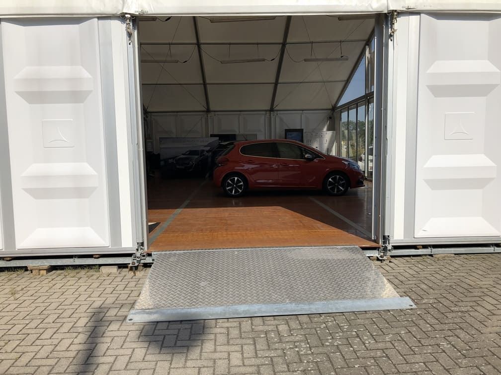 Une voiture rouge est garée à l'intérieur d'une grande tente blanche avec une rampe métallique. La porte de la tente est ouverte et le sol est en briques.
