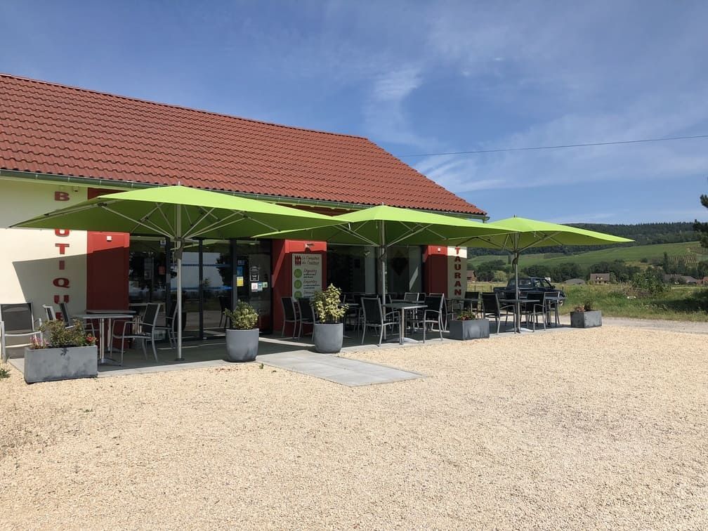 Un café avec une terrasse ombragée par des parasols verts. Le bâtiment a un toit rouge, un parking en gravier et un ciel ensoleillé.