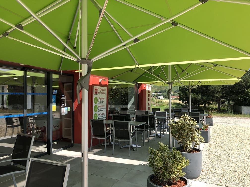 Café en plein air avec parasols verts, tables et chaises. Bâtiment aux portes et fenêtres vitrées.