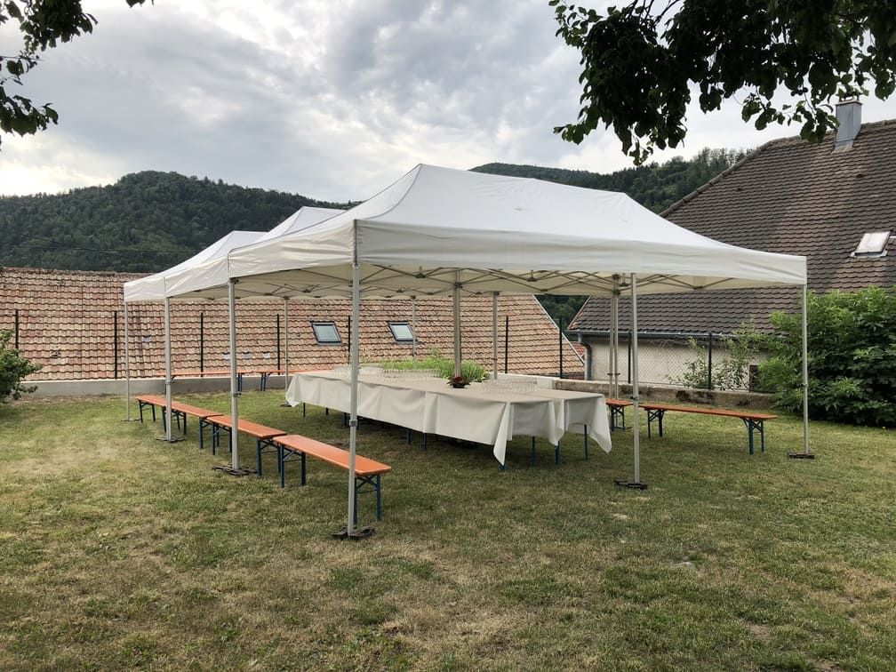 Longue table sous une tente blanche, avec des bancs, dans une cour herbeuse. Montagnes en arrière-plan.