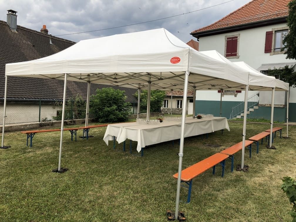 Des tentes blanches sont installées sur une pelouse, avec de longues tables et des bancs ; un bâtiment se dresse en arrière-plan.