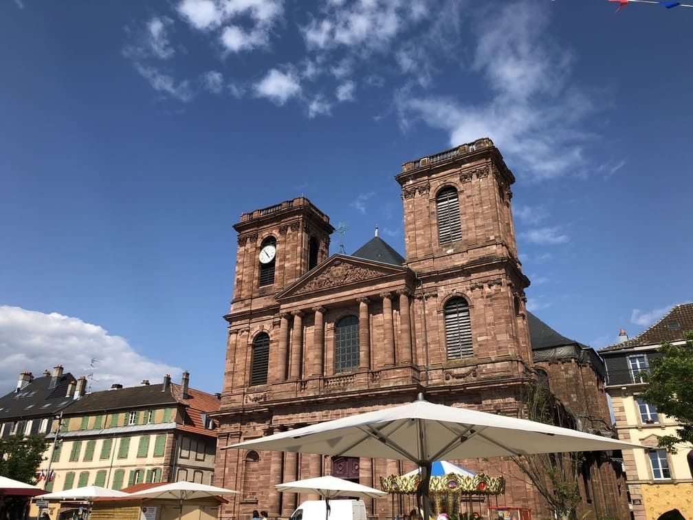 Église en briques rouges avec deux tours, une horloge et des parasols blancs se détachant sur un ciel bleu.